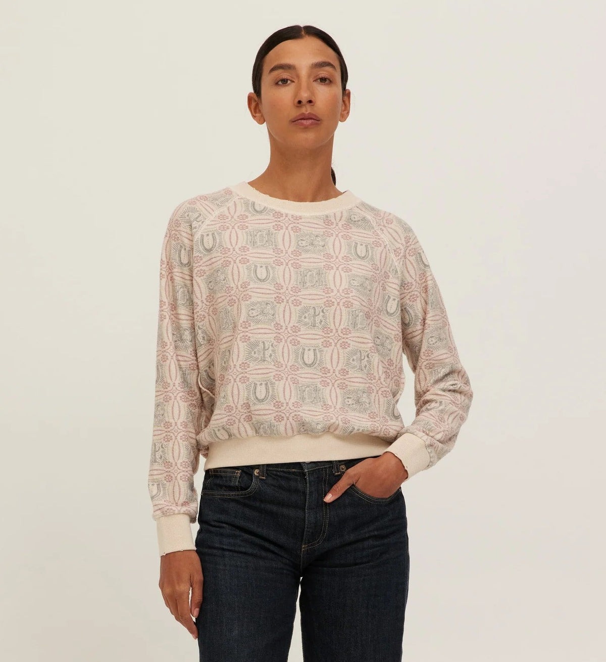 A model stands against a plain light background wearing a cream-colored crewneck sweatshirt with a geometric pattern, paired with dark blue jeans and dark brown shoes.