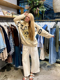 A woman posing in a clothing boutique wearing a beige graphic sweatshirt and cream wide-leg pants, surrounded by racks of clothes.