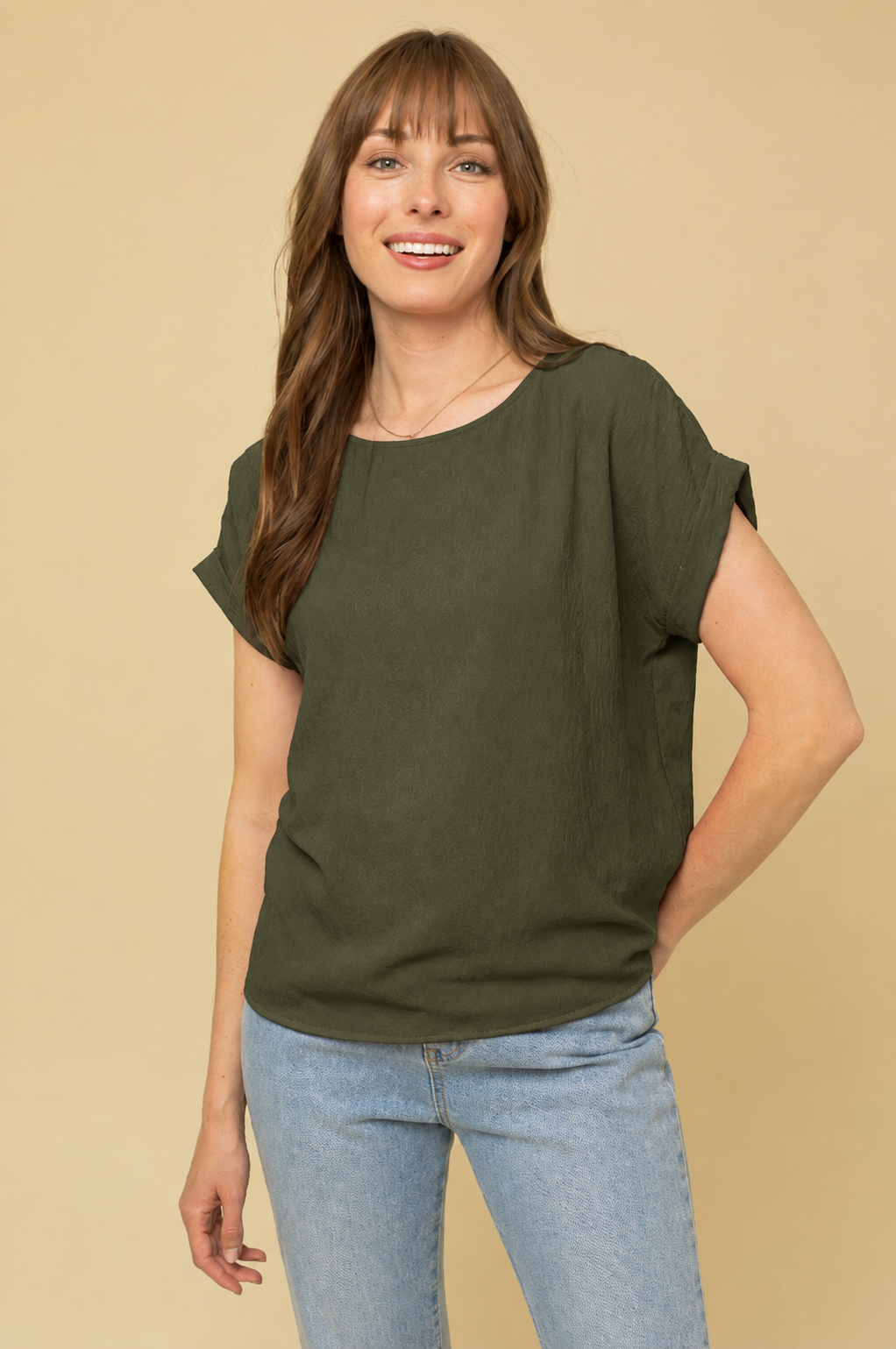 A woman wearing a short-sleeve olive green top and light blue jeans, standing against a solid beige background.