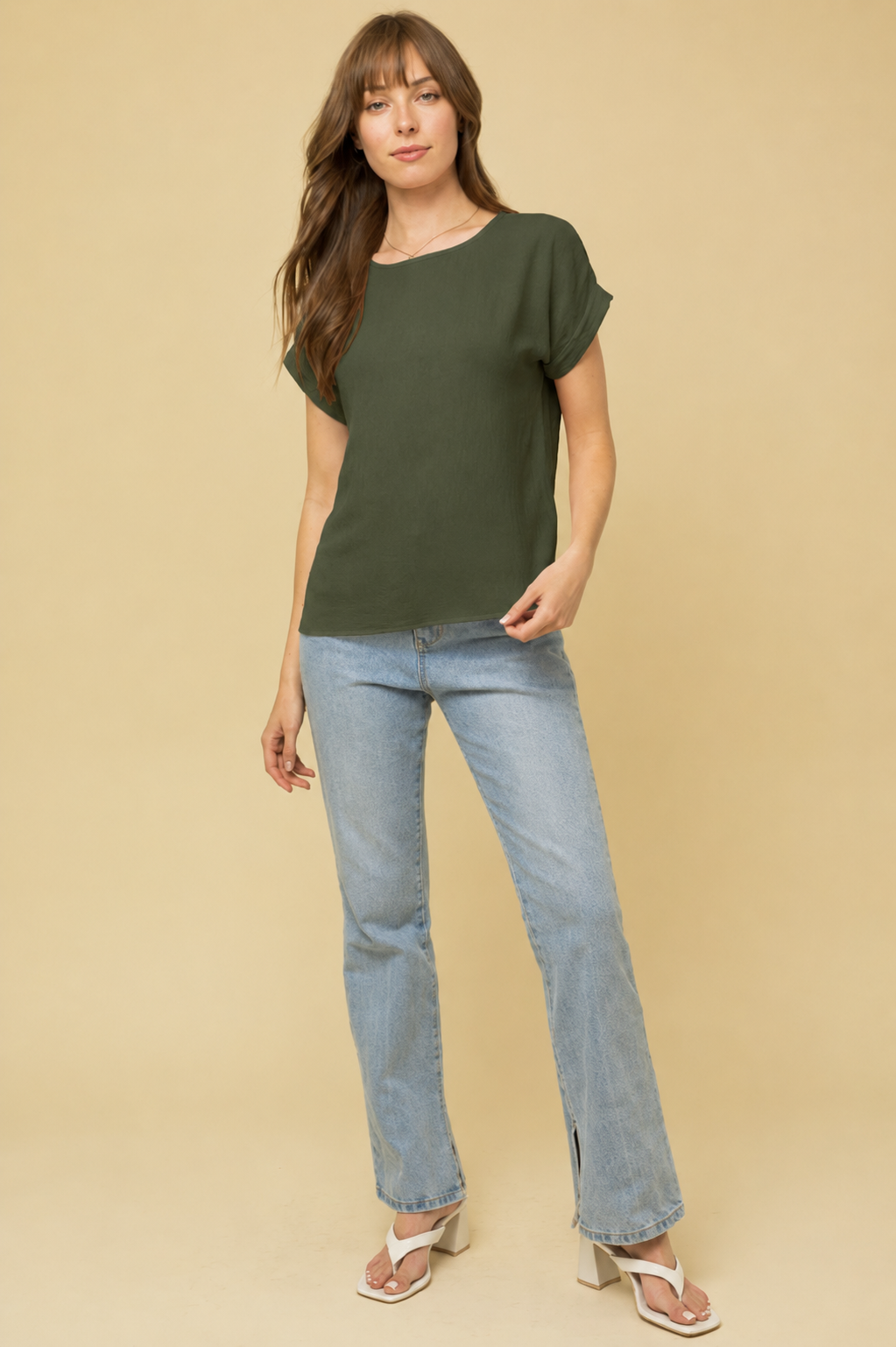 A woman modeling a dark olive green short-sleeve t-shirt and light-wash blue jeans against a solid beige studio background.