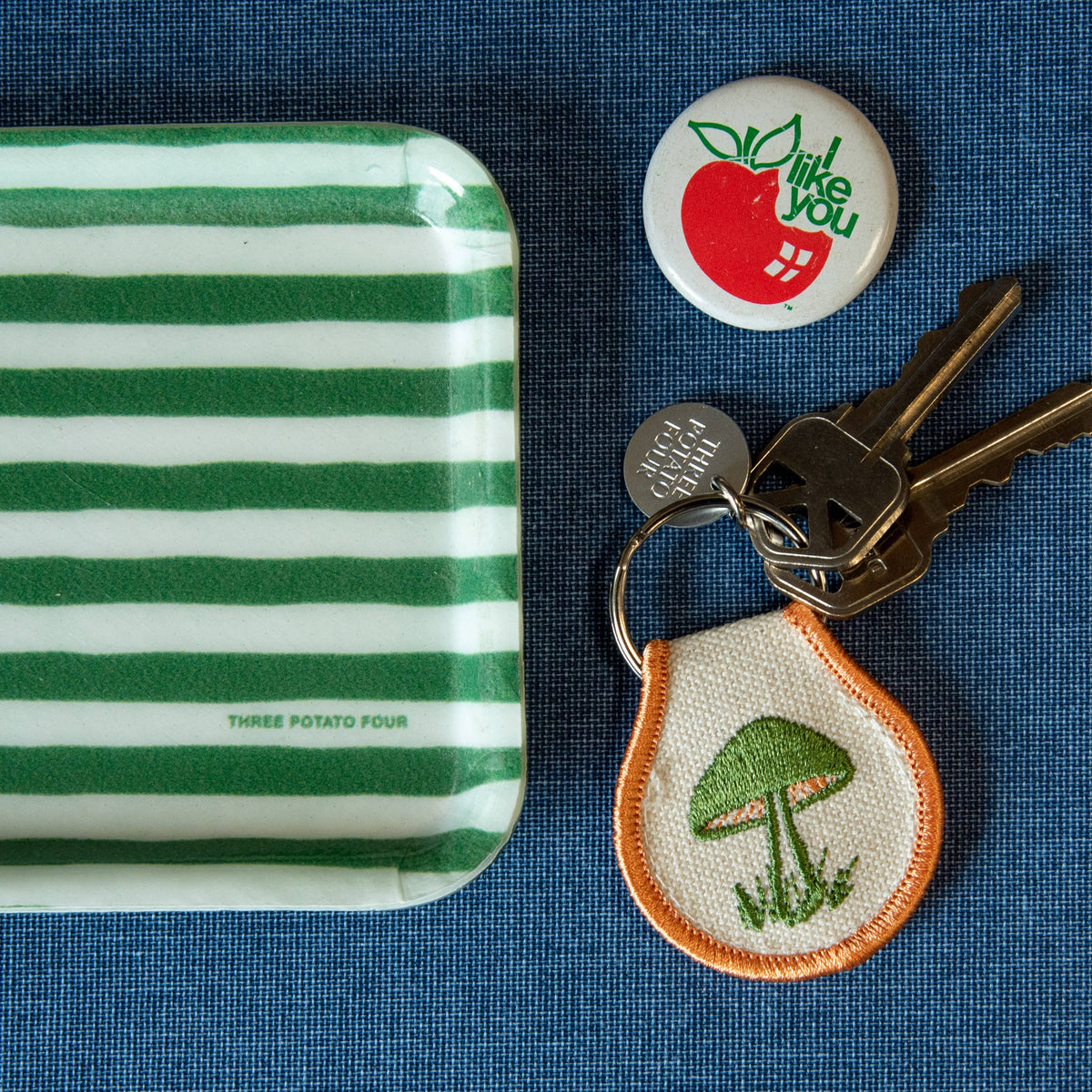 Striped container, button with apple design, keychain with mushroom, and keys on a blue textured background